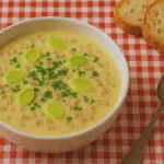 Suppenbowl mit Kartoffelsuppe, Hackfleischstückchen und Lauchscheiben auf einer rot-weiß karierten Tischdecke, Brotscheiben und ein Löffel komplettieren die Szene